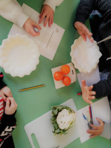 Bambini attorno ad un banco con piatti, tre mandarini al centro e un cavolfiore a metà